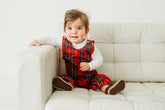 Baby wearing a red plaid outfit sitting on a white couch.
