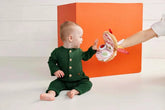 Baby in a green outfit playing with a toy in front of an orange box.