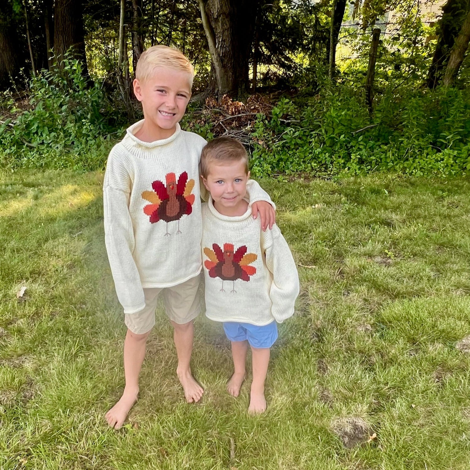 Two children wearing sweaters with turkey designs standing in a grassy area.