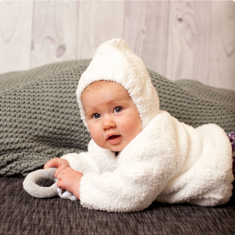 A baby wearing a fluffy white hooded jacket, lying on their stomach and holding a gray ring toy, set against a dark textured blanket and a light wood-paneled background.