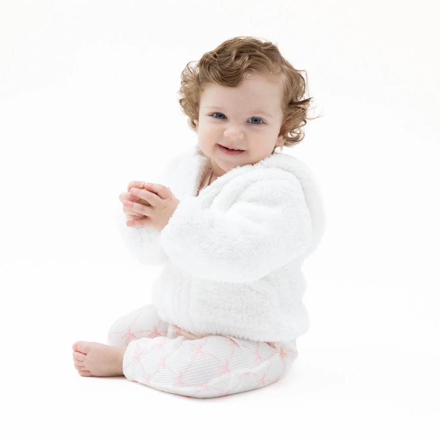 A smiling baby with curly light brown hair sits on a white studio background, wearing a plush white hooded robe and pale pink patterned pants.