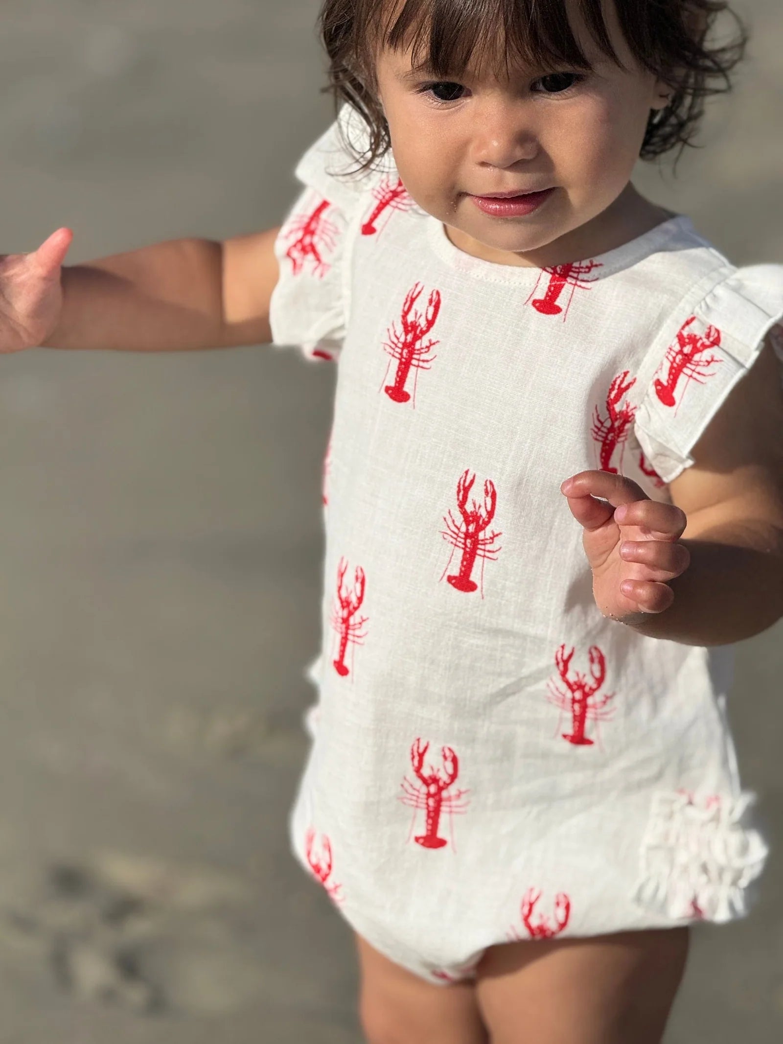 Child wearing a white outfit with red lobster patterns on a beach.