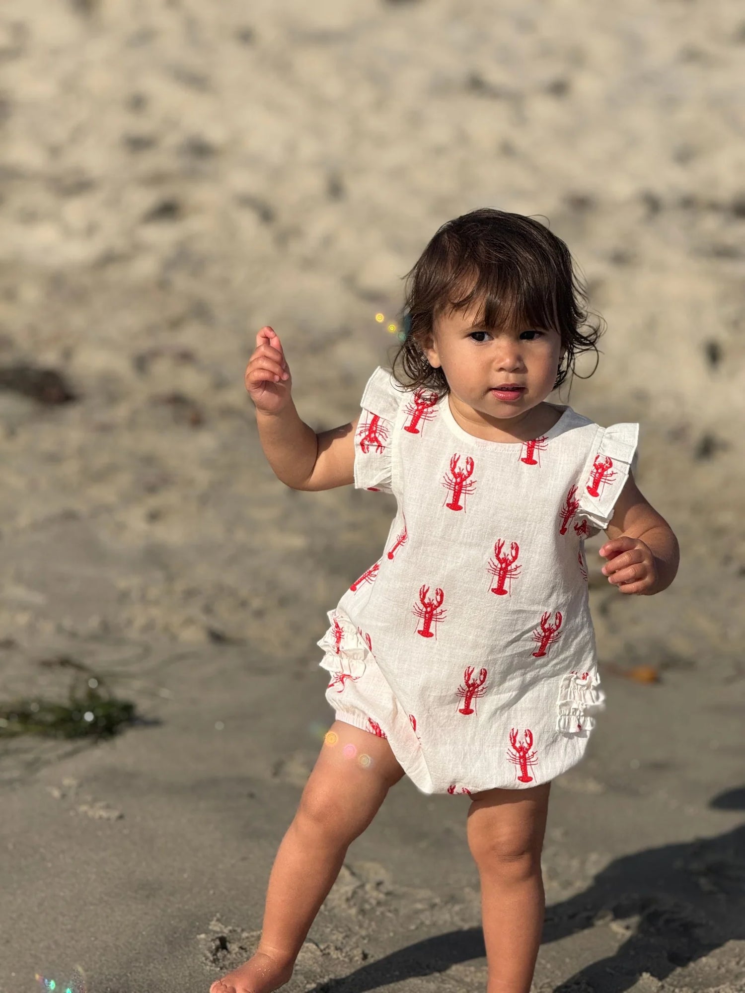 Child in a white dress with red lobster patterns on a sandy beach
