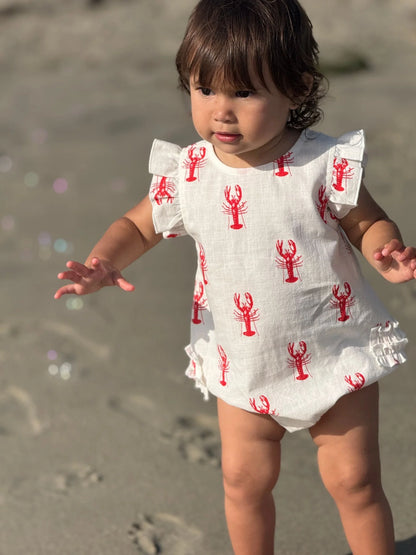 Child wearing a white romper with red lobster prints on a sandy beach.