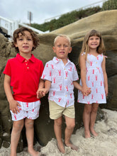 Three children standing on a sandy beach holding hands, wearing matching outfits with red and white patterns.