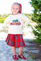 Young girl wearing a cream sweater with a Christmas design and a red plaid skirt outdoors.