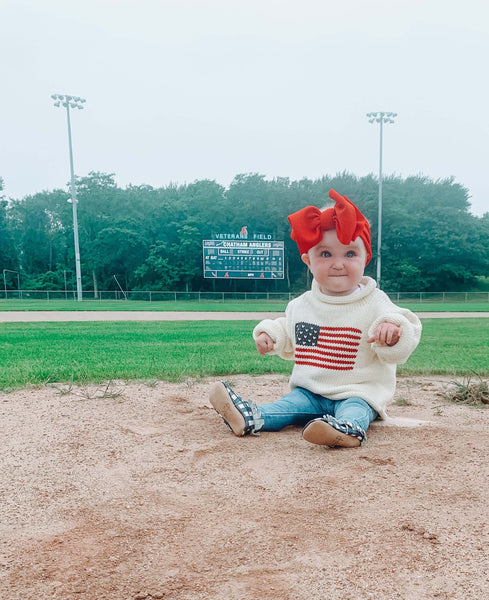 Ivory American Flag Roll Neck Sweater on Baby on baseball field, baby girl is wearing the sweater with jeans, gingham shoes and a red bow on head