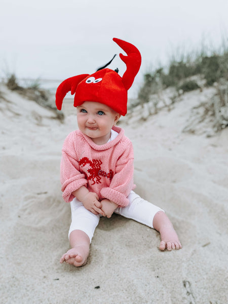 baby girl wearing Pink Lobster Roll Neck Sweater with white leggings and lobster claw hat on a beach